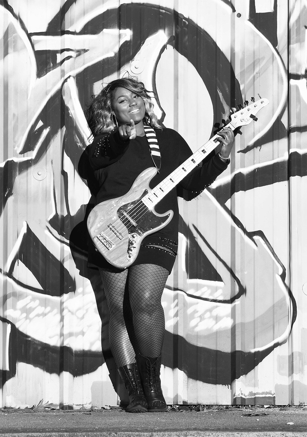 Téja Veal standing against a wall with street art holding a bass and pointing at the camera