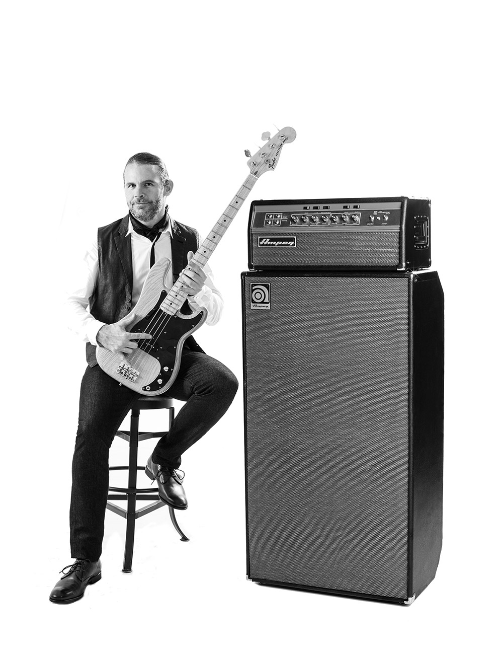 Tim Lefebvre sitting on a stool holding a bass next to an Ampeg stack